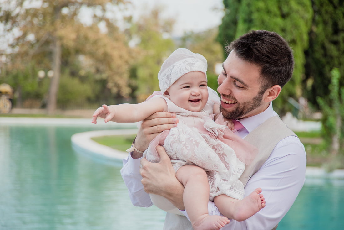 Φωτογραφία βάπτισης με το νονό να κρατάει το χαμογελαστό μωρό αγγαλιά μπροστά στο συντριβάνι του άλσους της Νέας Φιλαδέλφειας